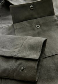 Camisa de manga larga con una suave y texturizada tela gris, que presenta puños con botones y cierres a presión, destacando una costura detallada y un acabado suave.