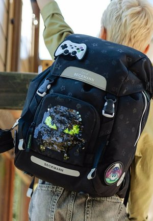Kind mit blondem Haar, das einen schwarzen Beckmann-Rucksack trägt, der mit einem Spielcontroller-Schlüsselanhänger und einem glitzernden Monster-Gesichtsfleck verziert ist.