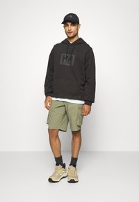 Haut à capuche marron foncé avec logo noir, shorts cargo vert olive et baskets beiges avec accents jaunes. Le modèle porte une casquette noire et des chaussettes grises.