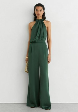 Mujer con un mono sin mangas verde oscuro con escote halter, sosteniendo un clutch dorado, de pie contra una pared blanca.