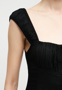 Κοντινό πλάνο ενός ατόμου που φοράει μαύρο φόρεμα με φαρδιές ραμμένες τιράντες και εφαρμοστό μπούστο.