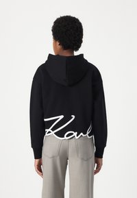 Schwarzer Hoodie mit einem weißen, geschwungenen "Karl"-Logo am Saum. Hergestellt aus weichem Stoff, hat er einen lässigen Schnitt und eine gefütterte Kapuze. Kombination mit hellgrauen Hosen.