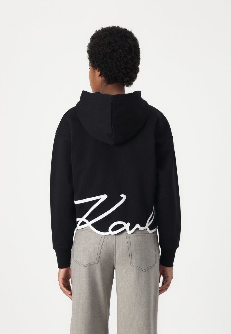 Schwarzer Hoodie mit einem weißen, geschwungenen "Karl"-Logo am Saum. Hergestellt aus weichem Stoff, hat er einen lässigen Schnitt und eine gefütterte Kapuze. Kombination mit hellgrauen Hosen.