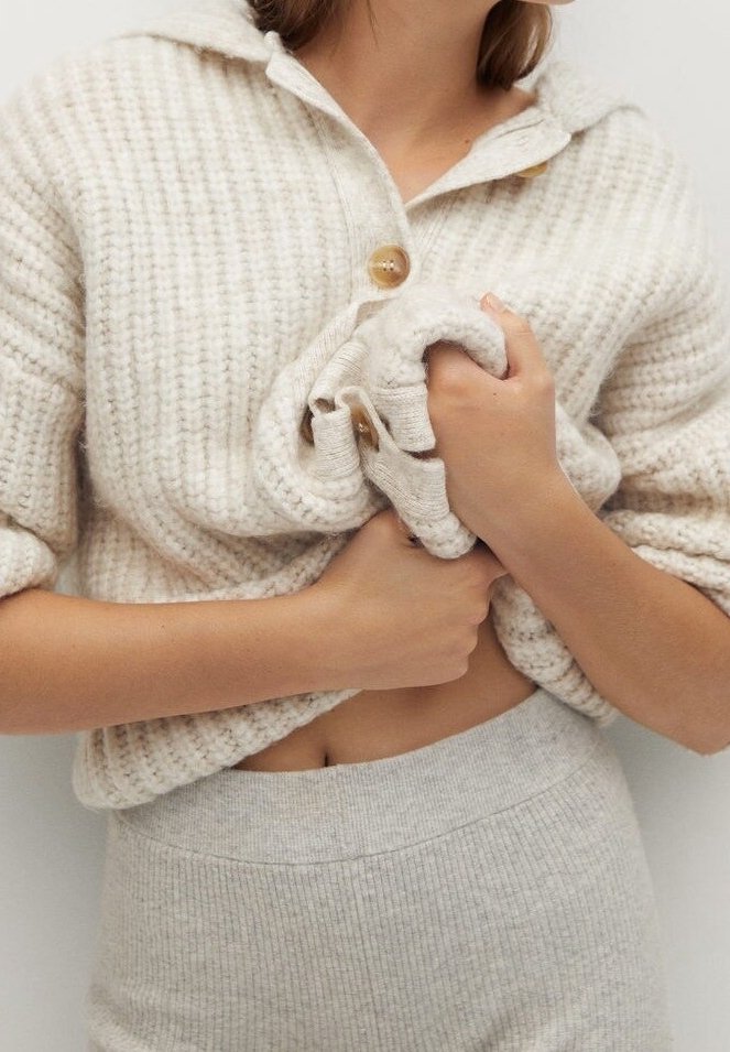 Femme portant et tenant un pull en tricot doux de couleur crème avec de gros boutons, assorti à une jupe ou un pantalon côtelé.