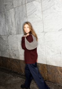 Frau mit langen Haaren, die einen bordeauxfarbenen Mantel und dunkelblauen Hosen trägt, eine graue, gewebte Umhängetasche hält und an einer mit Marmorfliesen verkleideten Wand entlanggeht.