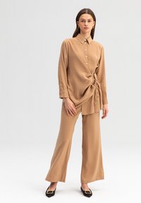 Camicia color beige con un dettaglio di cravatta in vita, dotata di chiusura a bottoni e maniche lunghe, abbinata a pantaloni a gamba larga dello stesso colore e scarpe nere.