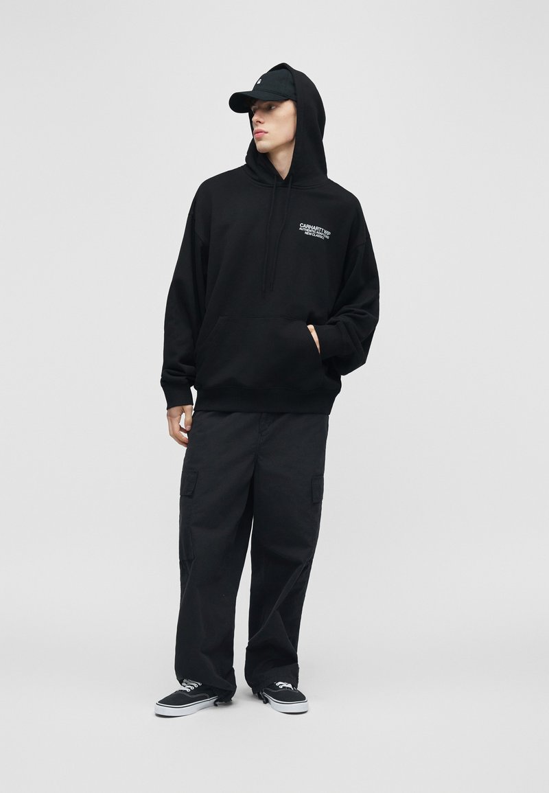 Giovane con felpa nera con scritta sul davanti, pantaloni cargo neri, cappellino nero e scarpe da ginnastica nere, in piedi davanti a uno sfondo grigio uniforme.