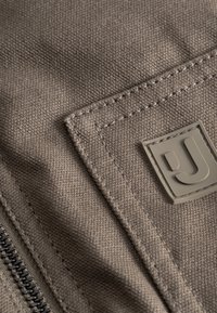 Sivá plátnová tkanina s textúrovaným povrchom, obsahujúca kontrastné šitie a gumový logopatch. Zahrnutý je viditeľný zips v čiernej farbe.