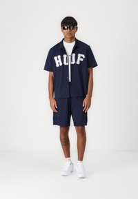 Marineblauw overhemd met korte mouwen en knoopsluiting, voorzien van een geperforeerde textuur en het "HUDF" logo, gecombineerd met bijpassende shorts en witte sneakers. Zwarte zonnebril gedragen.