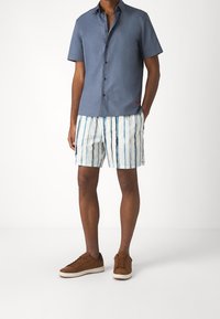 Blå skjorta med kort ärm och knappar, kombinerad med vita shorts med vertikala blå ränder och bruna mockaskor.