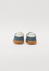 Jeans teniske z teksturiranim modrim zgornjim delom in belimi podrobnostmi. Imajo rjav gumijast podplat in oblazinjen hrbet za udobje.