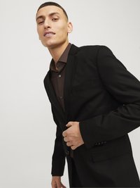 Black suit with a two-button closure, notch lapels, and a chest pocket. The model wears a brown shirt underneath. Smooth fabric texture.