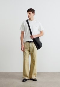 Νεαρός άνδρας που φοράει λευκό T-shirt, μπεζ παντελόνι με φαρδύ πόδι, μαύρα παπούτσια και μαύρη τσάντα χιαστί, στέκεται μπροστά σε λευκό, απλό τοίχο.