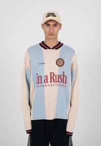 Homme portant un pull beige rayé bleu clair avec le texte "In a Rush International" et une casquette beige avec le texte "Always Late Club", immobile.