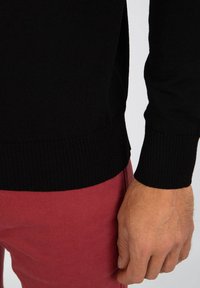 Zwarte gebreide trui met geribbelde manchetten en zoom, met een relaxte pasvorm. Gecombineerd met rode broek, gemaakt van een gestructureerde stof.