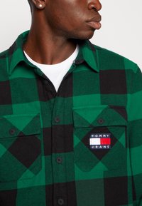 Camicia in flanella a quadri verde e nera con tasche sul petto con bottone e un patch del logo Tommy Jeans sulla tasca sinistra.