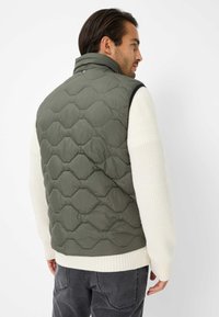 Grüne gepolsterte Weste mit hexagonalem Steppdesign, hohem Kragen und schwarzen Akzenten, getragen über einem strukturierten cremefarbenen Pullover, kombiniert mit grauen Hosen.
