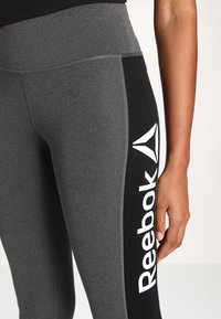Leggings atletici grigi con un pannello nero, dotati di un grande logo Reebok bianco. Realizzati in materiale elasticizzato, progettati per comfort e libertà di movimento.
