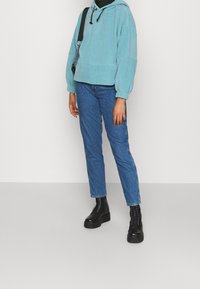 Persona che indossa un felpa con cappuccio turchese, jeans blu, stivali platform neri con lacci e borsa a tracolla nera, in piedi contro uno sfondo bianco semplice.