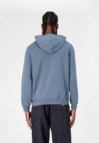 Sudadera de color azul claro con una gran capucha, puños y dobladillo acanalados. Tejido de algodón suave con un corte holgado. Presenta una sutil etiqueta con el logo en la cintura.