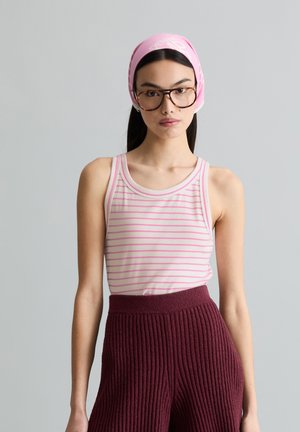 Young woman wearing brown-rimmed glasses, pink striped tank top, burgundy ribbed pants, and a pink headscarf, standing against a gray background.