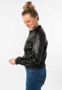 Zwarte leren bomberjack met een hoge kraag, geribbelde manchetten en taille. Voorzien van zijzakken en een gladde textuur. Draagt over denim.