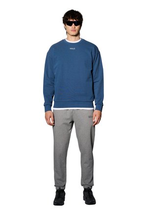 Blauwe sweatshirt met "HALO" logo, grijze trainingsbroek, zwarte sneakers en een zonnebril. Casual fit, katoen materiaal, ribgebreide manchetten en zoom.