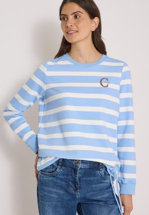 Kvinde iført en lyseblå og hvid stribet langærmet skjorte med et lille "Chase" logo, kombineret med blå jeans, der kigger til siden.