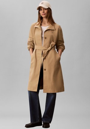 Frau trägt beigen Trenchcoat mit Gürtel, weißes T-Shirt, beige Kappe, dunkelblaue Jeans und dunkle Schuhe, steht mit den Händen in den Taschen.
