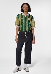 Grøn- og guldstribet kortærmet polo-shirt med struktureret stof, hvid krave og logo-badge, parret med sorte tapered bukser og sneakers.