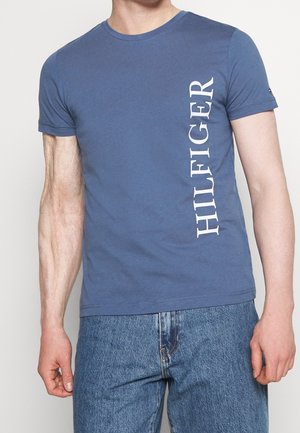 Niebieski bawełniany t-shirt z okrągłym dekoltem i krótkimi rękawami. Na lewej stronie biały, pionowy napis "HILFIGER". Prosty dół.