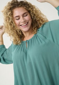 Blusa turchese con scollatura arricciata e dettaglio a oblò. Tessuto leggero e testurizzato con maniche lunghe. Design morbido e fluido.
