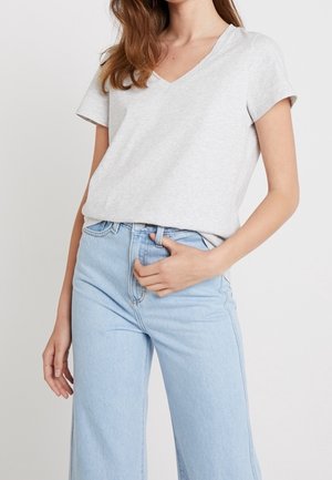 Kvinde iført en lysegrå T-shirt med V-hals og korte ærmer, stukket ned i højttaljede, lyseblå vidde jeans mod en ensfarvet baggrund.