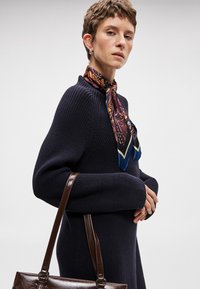 Ναυτικό πλεκτό φόρεμα με υψηλό γιακά, που διαθέτει μακριά μανίκια και άνετη γραμμή, συνδυασμένο με μια πολύχρωμη κεντημένη μαντήλι και μία καφέ τσάντα.