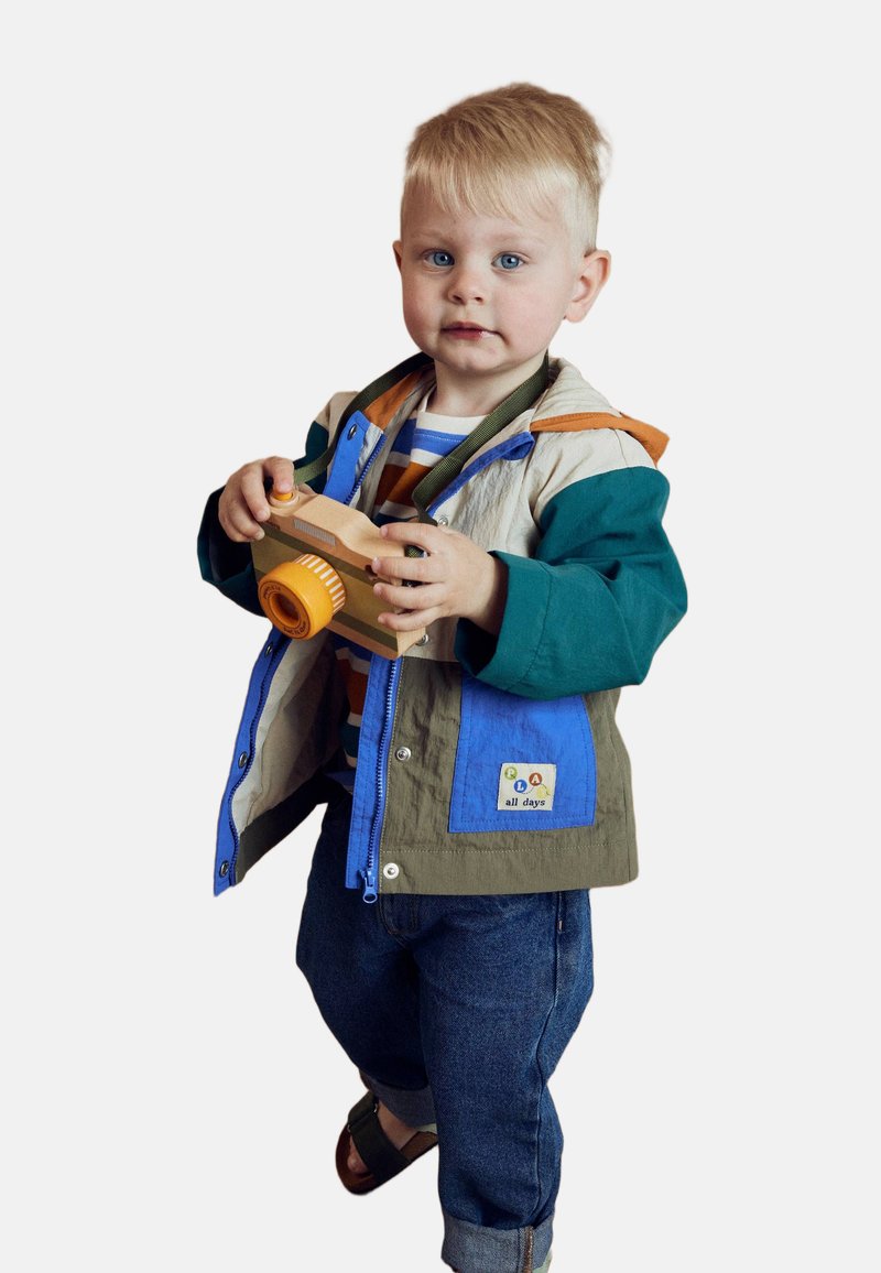 Enfant portant une veste légère multicolore avec des sections bleues, vertes et beiges, tenant un jouet caméscope en bois. Jeans en denim et sandales.