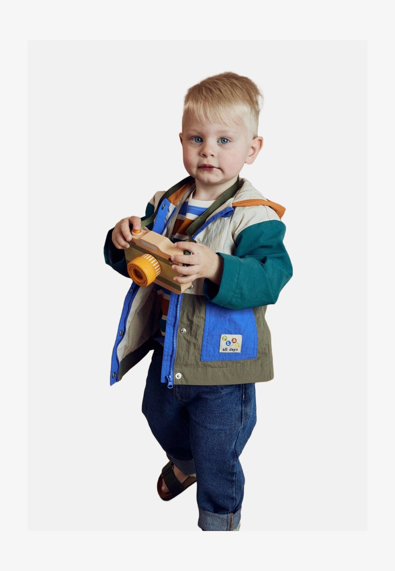 Enfant portant une veste légère multicolore avec des sections bleues, vertes et beiges, tenant un jouet caméscope en bois. Jeans en denim et sandales.