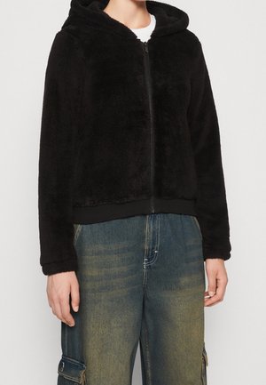 Czarna, puszysta bluza z kapturem i zamkiem z przodu, z prążkowanymi mankietami i dołem. Noszona z niebieskimi dżinsami w stylu cargo. Miękka tekstura i casualowy design.