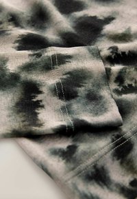 Bomuldsstof i grå og sort tie-dye mønster, blød tekstur med synlige syningsdetaljer langs kanten og manchetterne.