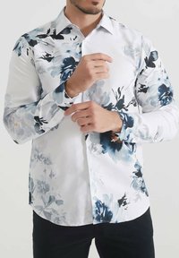 Chemise blanche avec un motif floral bleu, à boutons, à manches longues, texture lisse et accents bleus contrastants sur les poignets.