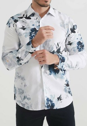 Chemise blanche avec un motif floral bleu, à boutons, à manches longues, texture lisse et accents bleus contrastants sur les poignets.