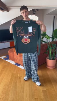 Young man in plaid pants holding dark green sweater with smiling reindeer and hanging ornaments in a modern living room.