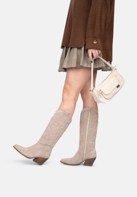 Botas de ante beige hasta la rodilla con punta afilada, costuras decorativas y tacón apilado. Llevadas con una falda de cuadros y un bolso ligero.