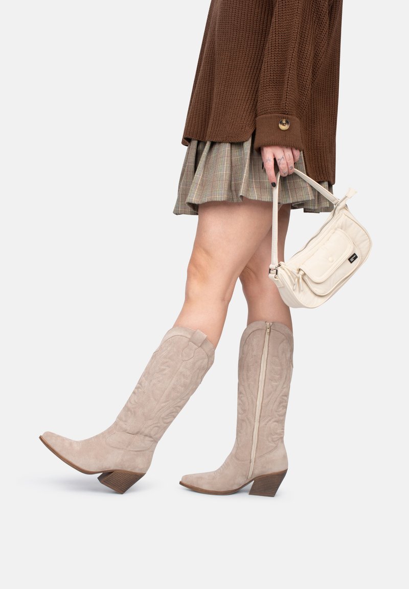 Botas de ante beige hasta la rodilla con punta afilada, costuras decorativas y tacón apilado. Llevadas con una falda de cuadros y un bolso ligero.
