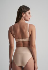 Donna di spalle che indossa biancheria intima seamless a vita alta color nude e un reggiseno con spalline regolabili, su uno sfondo grigio uniforme.