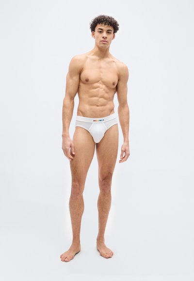 Νέος άνδρας που στέκεται ξυπόλυτος φορώντας λευκά εσώρουχα με το λογότυπο "DSQUARED2" στη ζώνη, σε απλό φωτεινό φόντο.