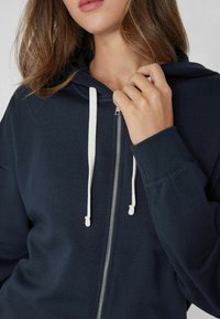 Marineblaues Zip-Hoodie mit Kapuze und Kordelzug. Ausgestattet mit einem silbernen Reißverschluss und gerippten Bündchen, gefertigt aus weichem Stoff mit glatter Oberfläche.