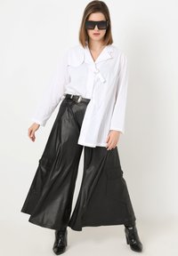 Weißes Langarmhemd mit spitzem Kragen, kombiniert mit schwarzen, weit geschnittenen Kunstlederhosen mit Seitentaschen und glänzender Textur. Schwarze Sonnenbrille.