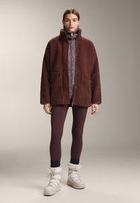 Hnědá fleecová bunda s vysokým límcem přes vzorovaný fialový svetr, doplněná vínovými legínami a bílými zateplenými botami.