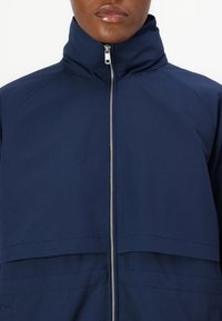 Veste coupe-vent bleu marine avec col montant, fermeture zippée avant et coutures horizontales sur la poitrine. Tissu lisse et léger.