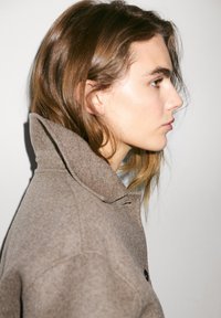 Ung person i profil med lys hud og brunt hår iført en højhalset, brun uldfrakke mod en ensfarvet lys baggrund.